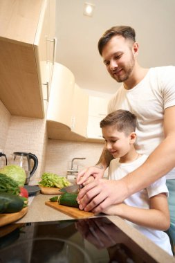 Erkek ebeveyn oğlanı bıçakla alıp mutfakta nasıl salatalık kesileceğini gösteriyor.