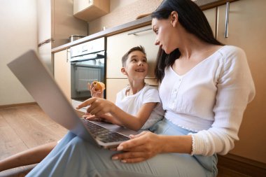 Mutfak zemininde sorusu olan annesine dönen, ısırılmış elmalı meraklı bir çocuk.