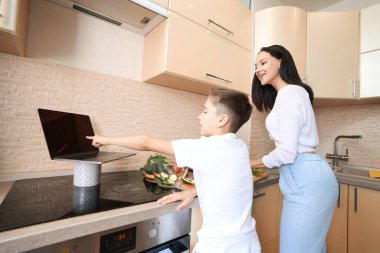 Çocuk, mutfakta yemek pişirmede annesine yardım ederken bilgisayar ekranını işaret ediyor.