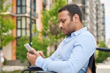 Serious focused disabled male person sitting in wheelchair and reading Internet news on mobile phone