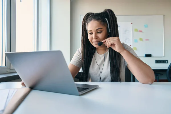 Smiling focused female office worker in headset sitting at laptop during video call