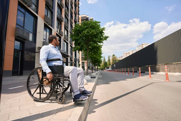 Disabled person sitting in manual wheelchair on sidewalk curb and looking aside