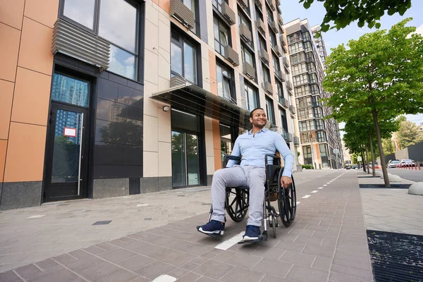 Smiling happy disabled person riding his wheelchair past office building on pavement