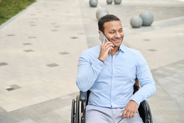 Joyous disabled person sitting in wheelchair with mobile phone pressed to ear