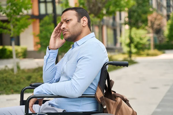Sad lonely young man seated in wheelchair on sidewalk suffering from headache