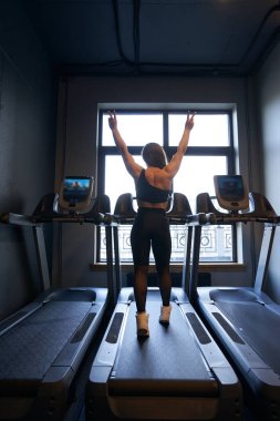 Koşu bandında yürürken iki eliyle zafer işareti gösteren atletik bayanın arka görüntüsü