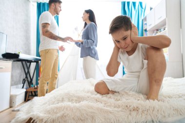 Çocuk kulaklarını kapatmış, ebeveynlerinin kavgalarını duymak istemiyor, ki bu da çocuk odasında oluyor.