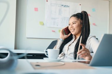 Smiling female entrepreneur sitting at office desk and looking into distance during phone talk
