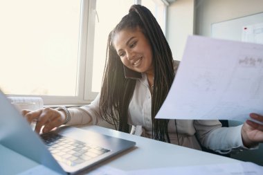 Smiling female engineer with technical drawing in hand looking at laptop screen during phone talk