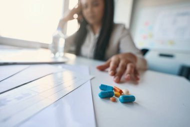 Kadın ofis çalışanının masamda oturup ilaçlara uzanırken çekilmiş fotoğrafı.