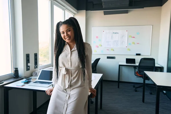 Smiling happy corporate employee sitting on edge of table and looking before her