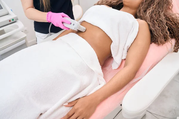 Specialist-cosmetologist in a modern salon of aesthetic medicine performs the procedure of hardware impact of smas-lifting on the abdominal area