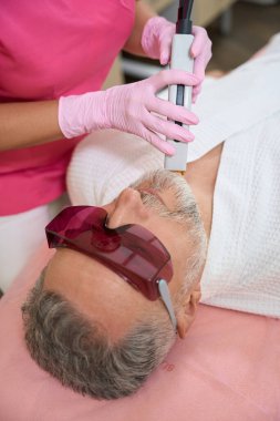 Gözlüklü sakallı adam yüzüne lazer tüyü aldırıyor. Pembe eldivenli kadın doktor.