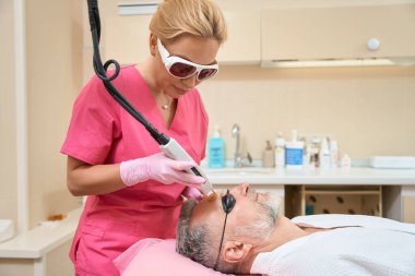 Female cosmetologist in glasses performs laser hair removal to adult man with a beard