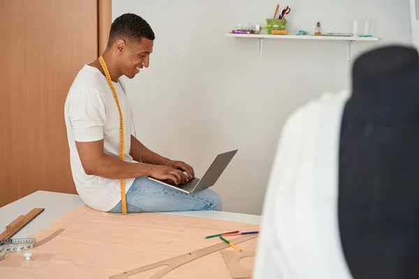 Happy young man is sitting on desk with drawings and typing on notebook while sewing