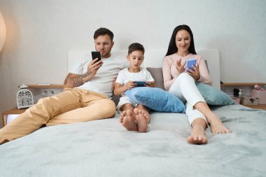 Baba, anne ve oğul rahat bir şekilde yastıkların arasında, akıllı telefonlarıyla yattılar.