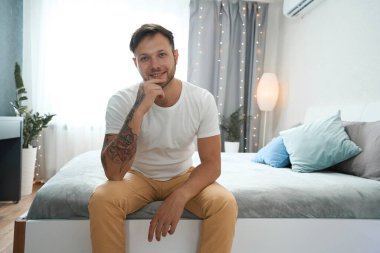 Yüzünde gülümseme olan neşeli genç adam yatak odasında yumuşak bir yatakta oturuyor, sakin ve mutlu.