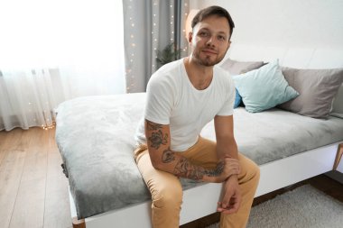 Gülümseyen genç adam yatak odasındaki yatağın üzerinde oturuyor, oda temiz ve rahat.