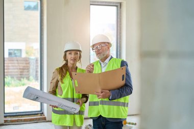 Realtor with drawings, and a foreman with folders in their hands are talking, standing in an unfinished house