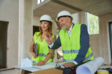 Smiling realtor and foreman inspecting house while standing near table with blueprints