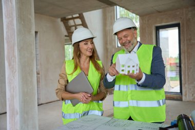 Smiling adult foreman holding model of house in his hand and examining it together with real estate agent