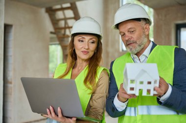 Agent holds open laptop in hands and looks at model of house, which shows gray-haired male foreman in protective helmet