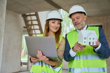 Realtor holds open laptop in hands and looks at model of house, which shows foreman in in costume and protective helmet