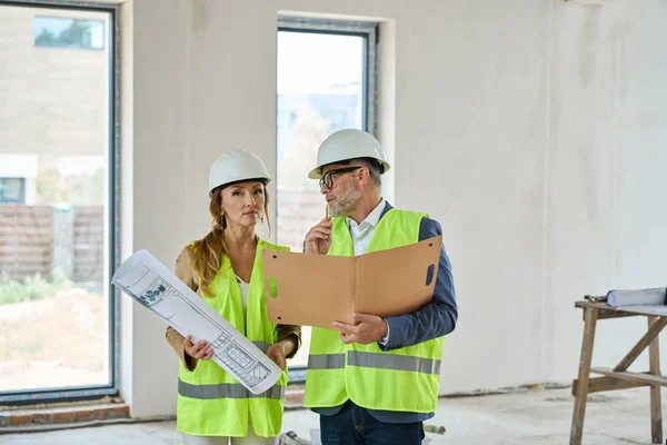 Real estate agent with blueprints and a foreman with folders in their hands are talking while standing in an unfinished house