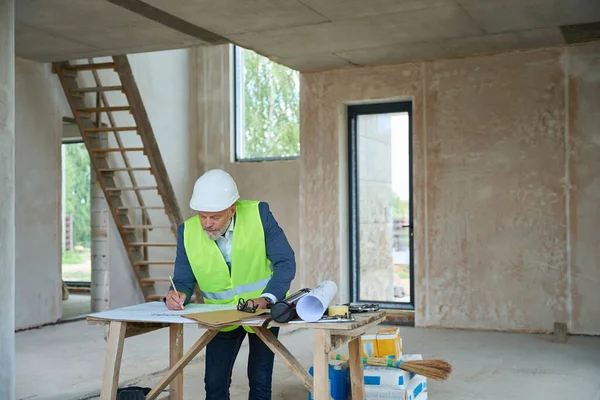 Foreman in protective helmet writes on drawings, which are laid out on table. Against background of concrete walls and staircase