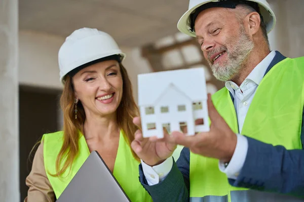Happy foreman in safety helmet holds small model of house in his hand and shows it to real estate agent