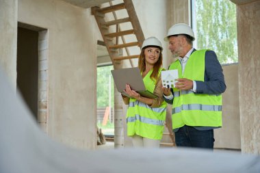 Smiling woman real estate agent and foreman looking at laptop. Man holding a model of house for construction