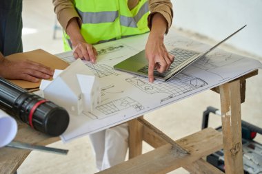 Close-up of hand of man and woman, which points to drawings lying on table near laptop. Couple at construction site