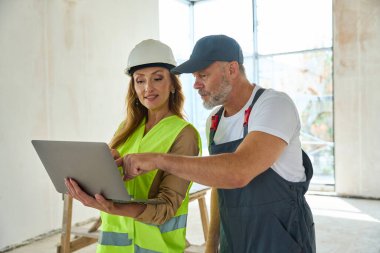 Gray-haired builder points a finger at open laptop held by pretty sales manager. Man and woman standing at construction site