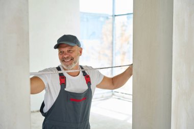 Joyful gray-haired builder measures width of the doorway with tape measure. Photo of man working on construction site
