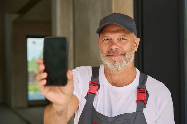 Взрослый Строитель Комбинезоне Бейсболке Показывает Телефон Мужчина Стоит Перед Домом — стоковое фото