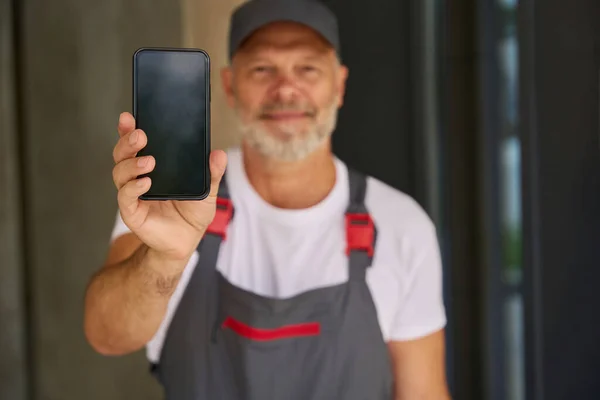 Улыбающийся Строитель Кепке Комбинезоне Показывает Мобильный Телефон Человек Стоит Перед — стоковое фото