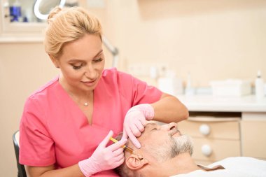 Beautiful female cosmetologist makes a beauty injection to a bearded man near the ear