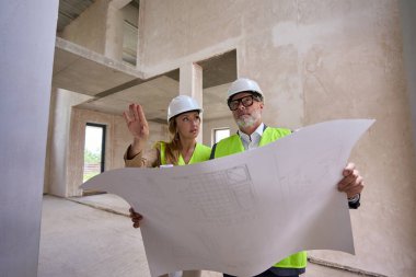 Foreman in glasses opened drawing and discusses it with realtor. They stand inside unfinished house in protective helmets