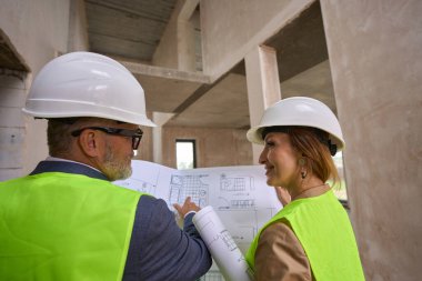 Foreman with beard is discussing open drawing of house with handsome manager. Woman holds second folded drawing in her hands