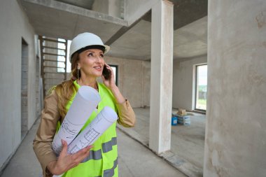 Smiling female realtor talking on mobile phone and holding rolled up blueprints of new houses