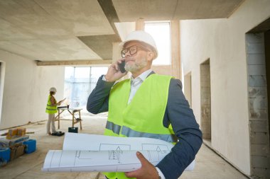 Foreman holds folded drawings in his hands and speaks on a mobile phone while standing in an unfinished house