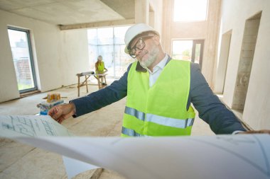 Male foreman holds expanded drawing of house in his hands and speaks on mobile phone. He stands inside unfinished house