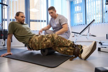 Patient in camouflage uniform does special rehabilitation exercises under the supervision of qualified instructor, he uses knee roll and weights clipart