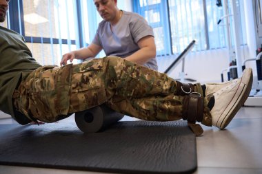 Military patient does rehabilitation exercises under the supervision of a physiotherapist, he uses a special popliteal roller and fixation tourniquet