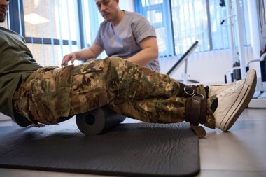 Military patient does rehabilitation exercises under the supervision of a physiotherapist, he uses a special popliteal roller and fixation tourniquet clipart