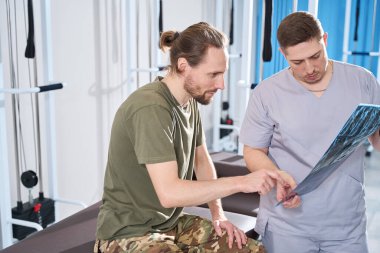 Soldier in military clothes and his attending physician communicate in a rehabilitation room and look at x-rays