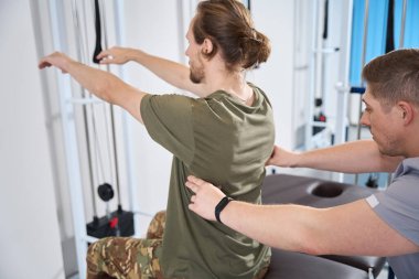 Bearded military man in camouflage pants undergoes session of physiotherapy exercises under supervision of doctor of physiotherapist in military hospital clipart