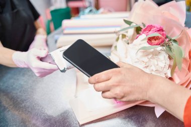 Woman hand holds phone near terminal to pay for ordered bouquet in flower boutique. Nearby lies ready-made bouquet of flowers.