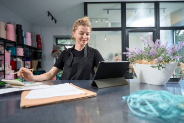Smiling female florist looking at tablet and counting sales, orders sitting at counter of her flower shop and checking documents