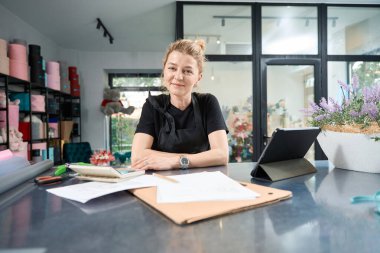 Cute florist sits at table of his own floral store and smiles sweetly at camera. Nearby are documents, calculator, tablet